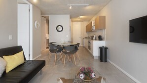 Un salon moderne avec un canapé noir, une table à manger en verre entourée de quatre chaises, une petite table basse agrémentée d’objets décoratifs, un coin cuisine avec des placards et des appareils électroménagers, et un téléviseur fixé au mur.