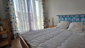 A bedroom with a bed, a chair, a nightstand, a lamp, and a window with curtains. - Beautiful, bright apartment at Les Sables Blancs in Concarneau - 150m from the beach and the GR34 (Concarneau)