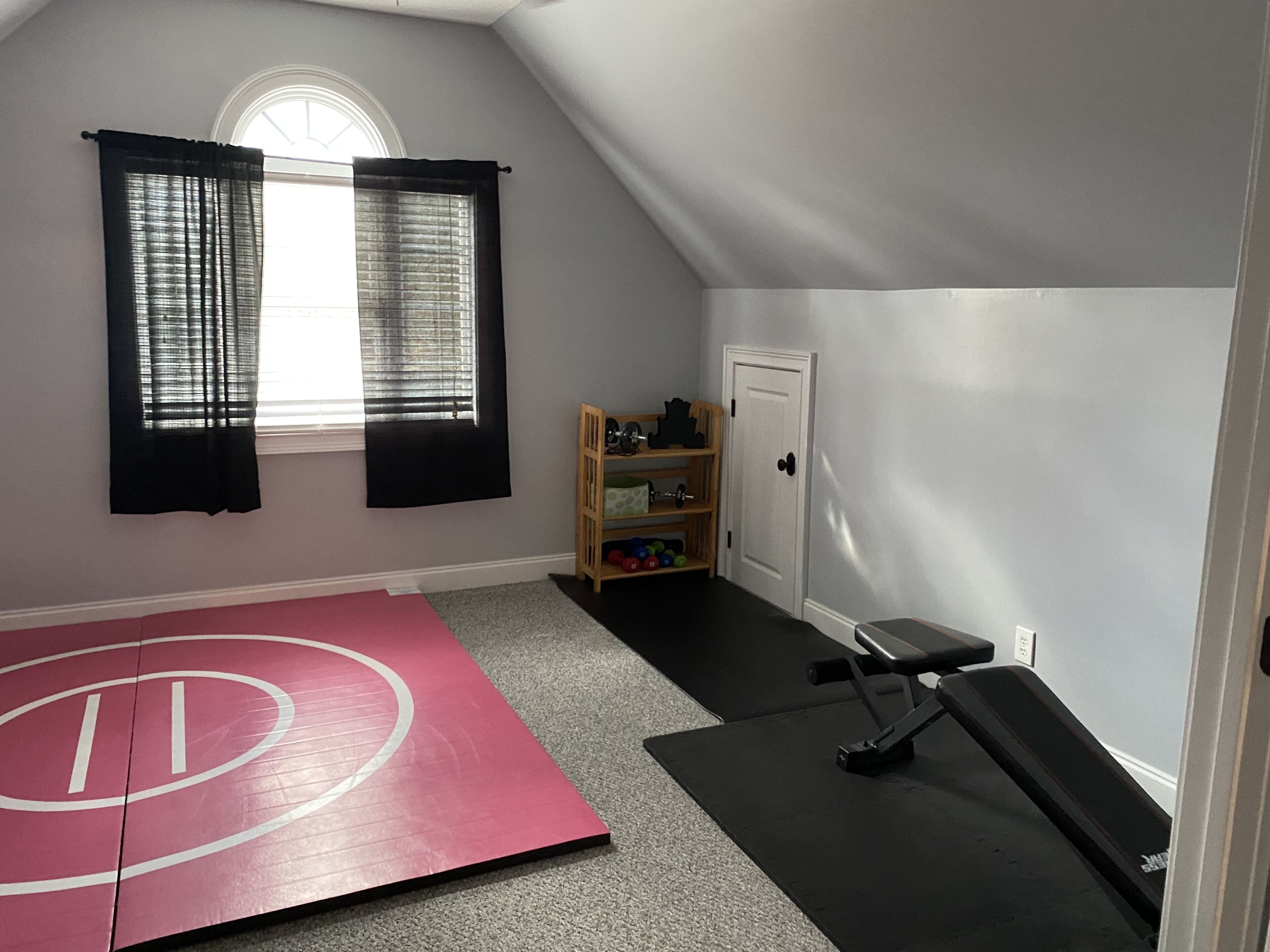 A small, well-lit room with a sloped ceiling, a red exercise mat, a black exercise bench, a wooden shelf with toys, and a door.