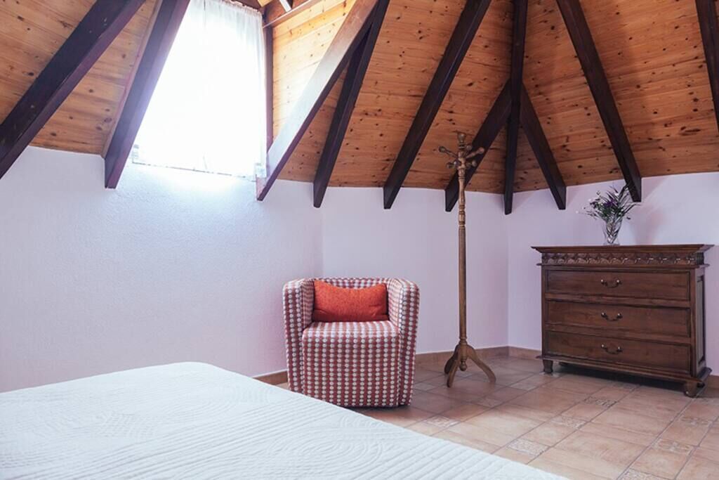 Un dormitorio con techo de madera inclinado, un sillón a cuadros, un aparador de madera y una cama.