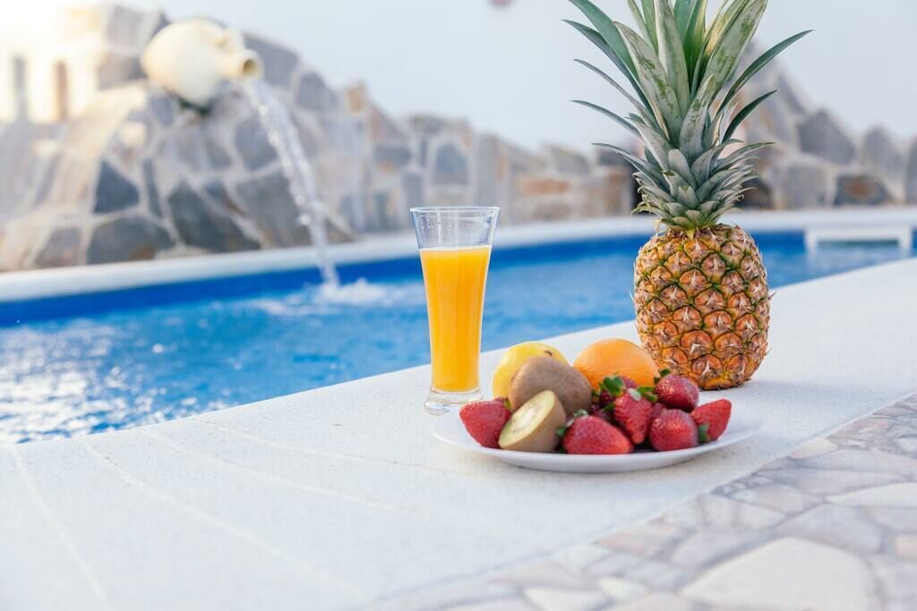 Una mesa junto a la piscina con un vaso de jugo de naranja, una piña, kiwis, fresas y naranjas.
