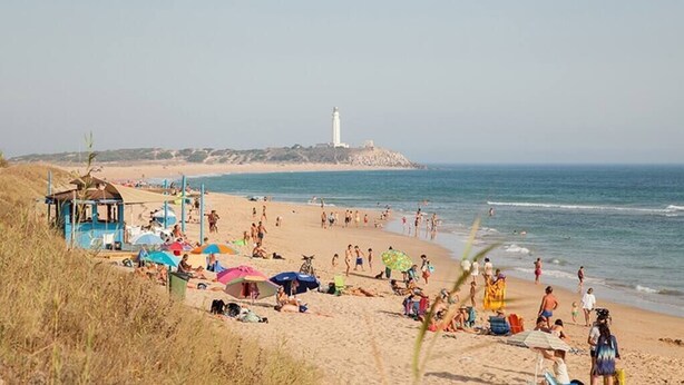 Una playa con un faro al fondo, gente tomando el sol y sombrillas de playa.