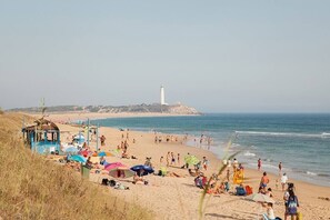 Uma praia com um farol ao fundo, pessoas tomando sol e guarda-sóis.