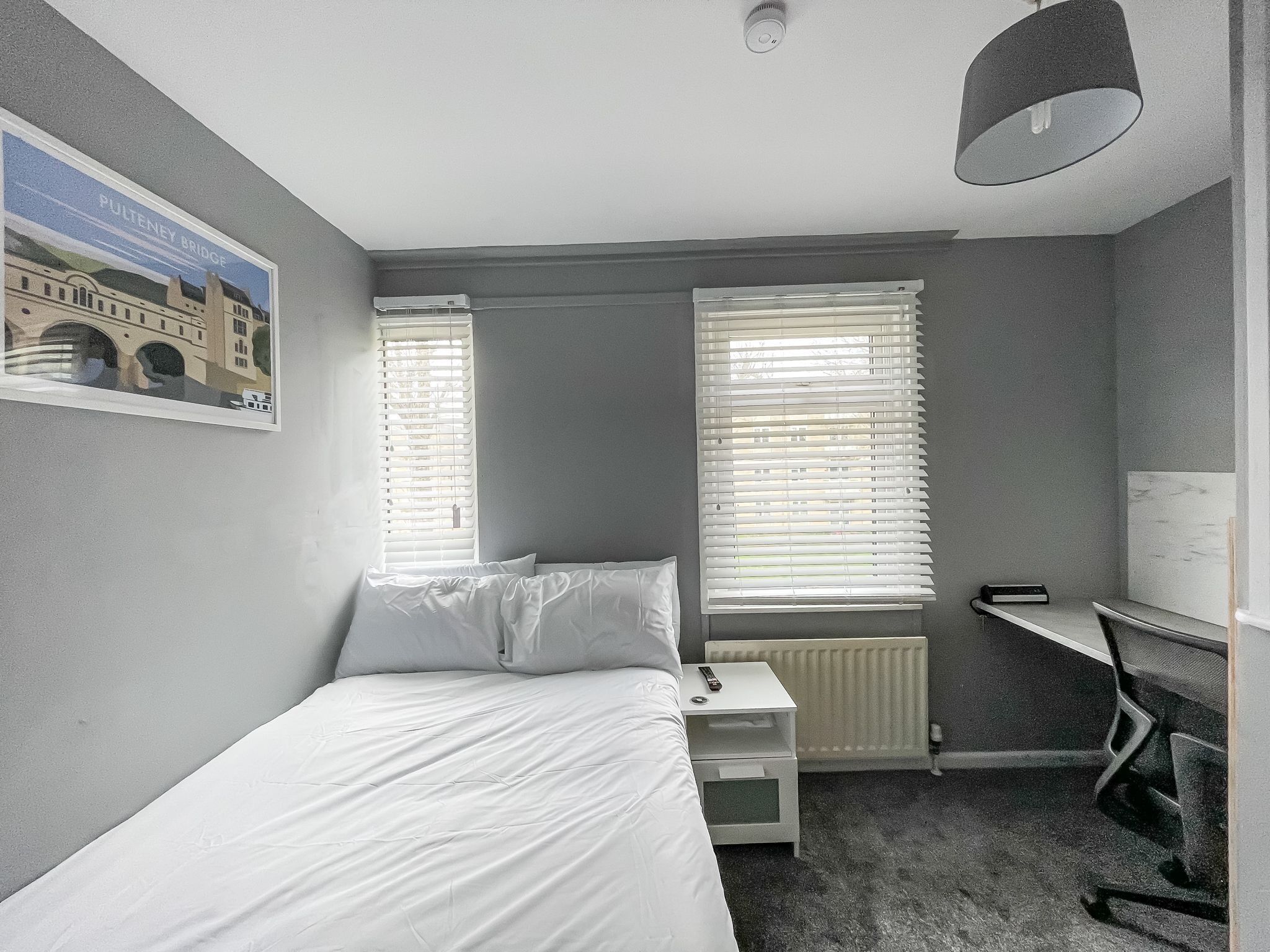 A hotel room with a bed, a desk, and two windows with blinds.