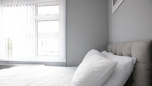 Een net opgemaakt bed met witte beddengoed en kussens, een grijs gestoffeerd hoofdeinde, en een raam met zonwering.