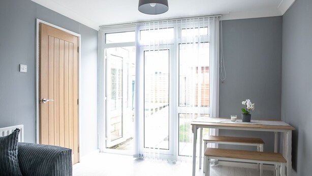 Een lichte kamer met een houten deur, een witte tafel met bankjes en uitzicht door grote ramen.