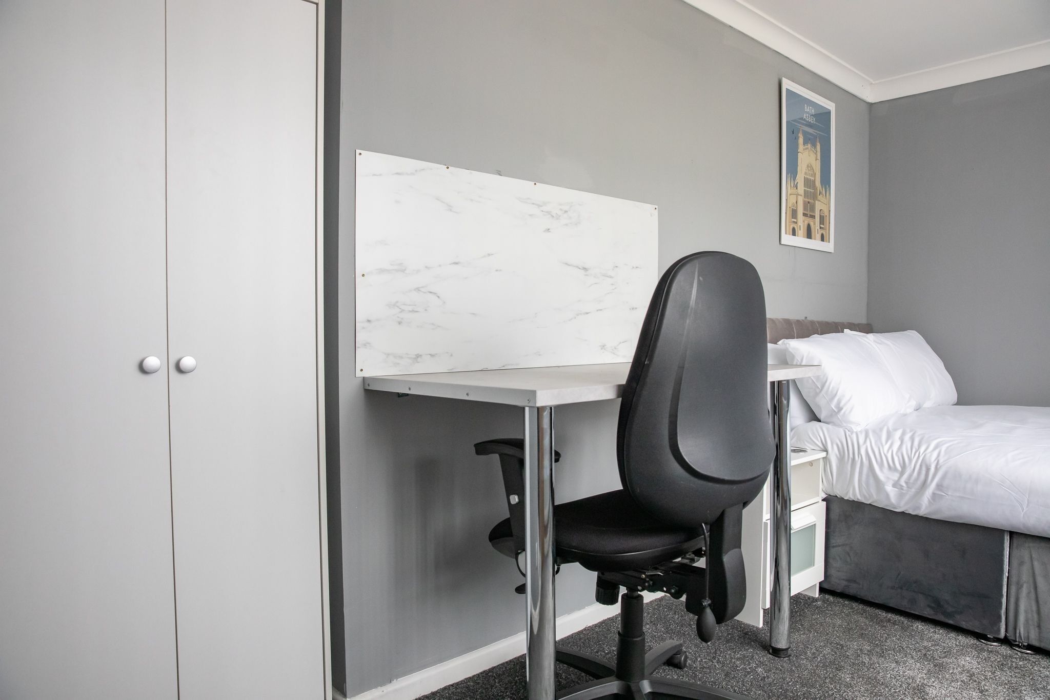 A bedroom with a bed, a desk with a chair, a whiteboard, and a framed picture on the wall.