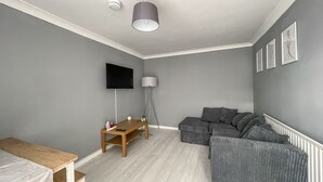 Een moderne, goed verlichte kamer met grijze wanden, een houten tafel, een grijze bank en een aan de muur bevestigde televisie.