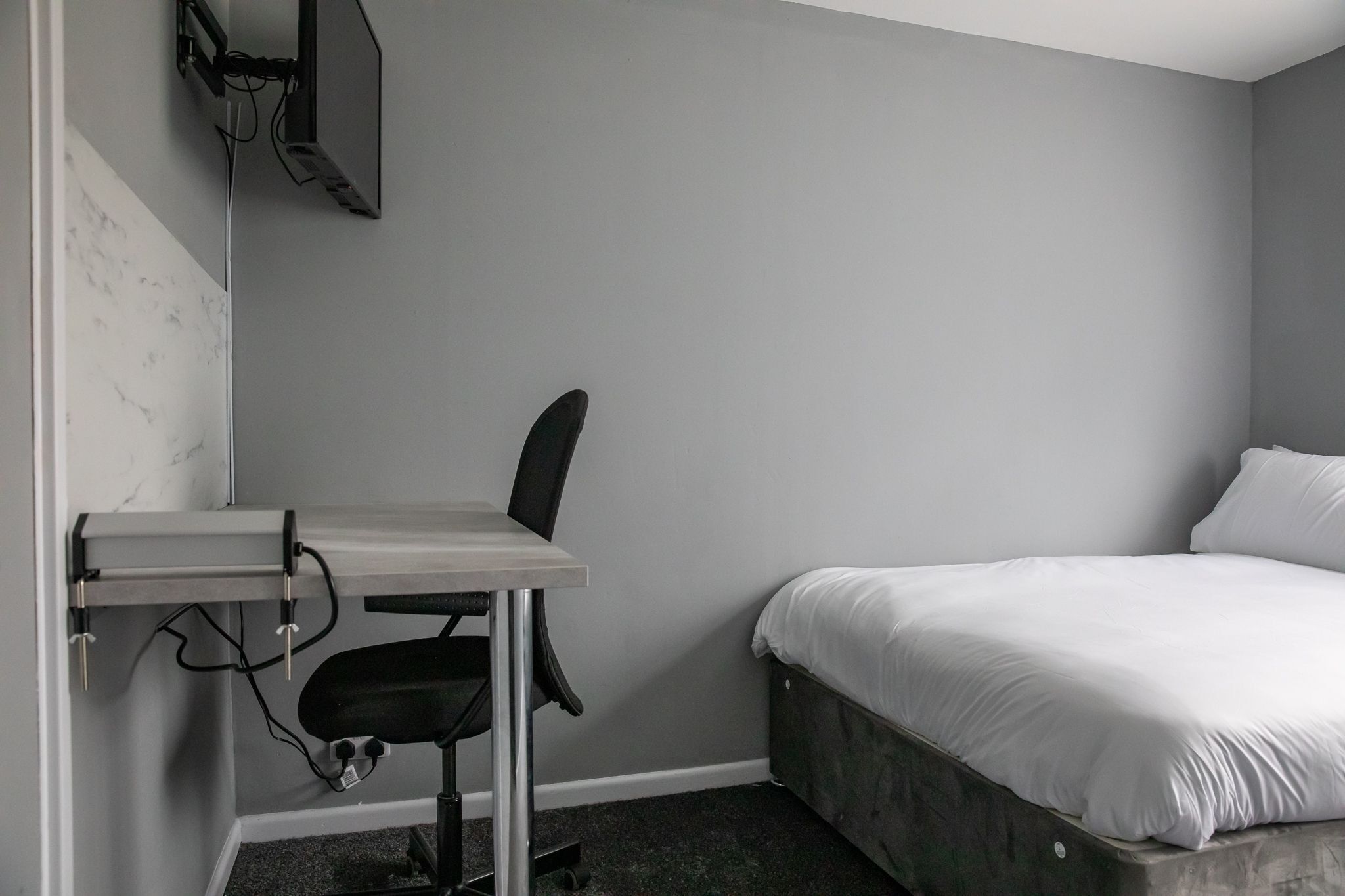 A small, modern bedroom with a single bed, a desk with a chair, and a mounted TV.