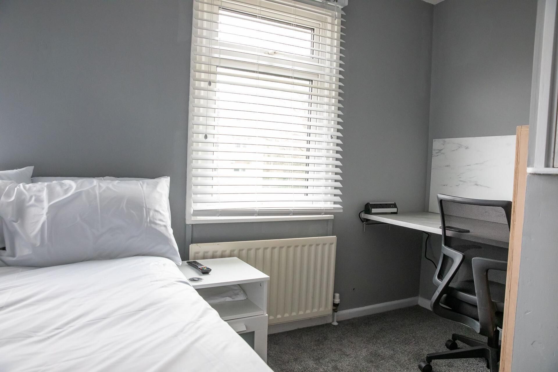 Une chambre à coucher comprenant un lit, un bureau, une chaise et une fenêtre avec des stores.