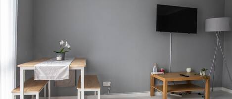 A room with a grey wall, a mounted TV, a wooden table with a white tablecloth, a bench, and a small shelf with a plant.