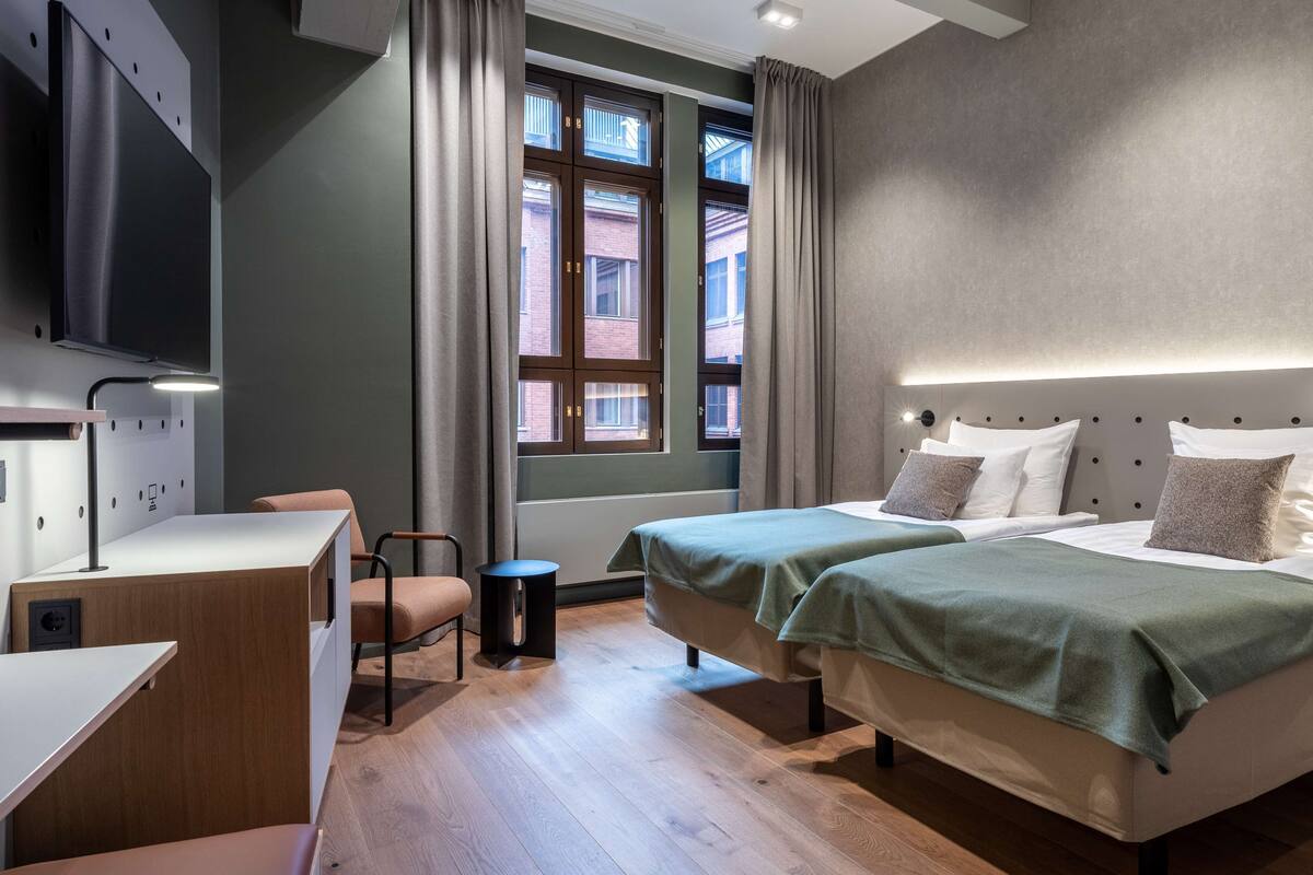 Une chambre d’hôtel moderne avec deux lits, un bureau, une chaise et une grande fenêtre avec des rideaux.