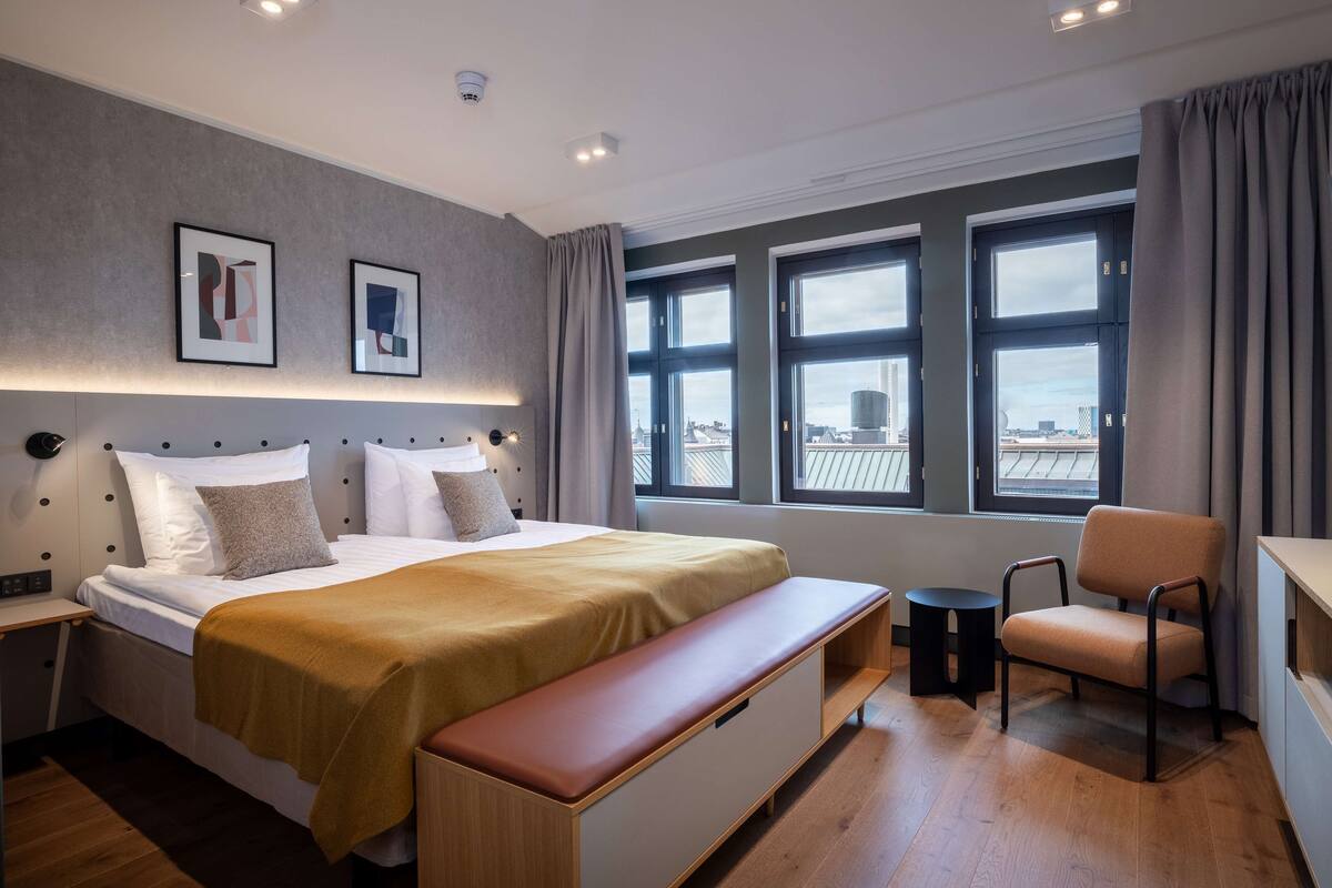 Une chambre d’hôtel moderne avec un grand lit, une banquette, une chaise et une vue sur la ville grâce à de grandes fenêtres.
