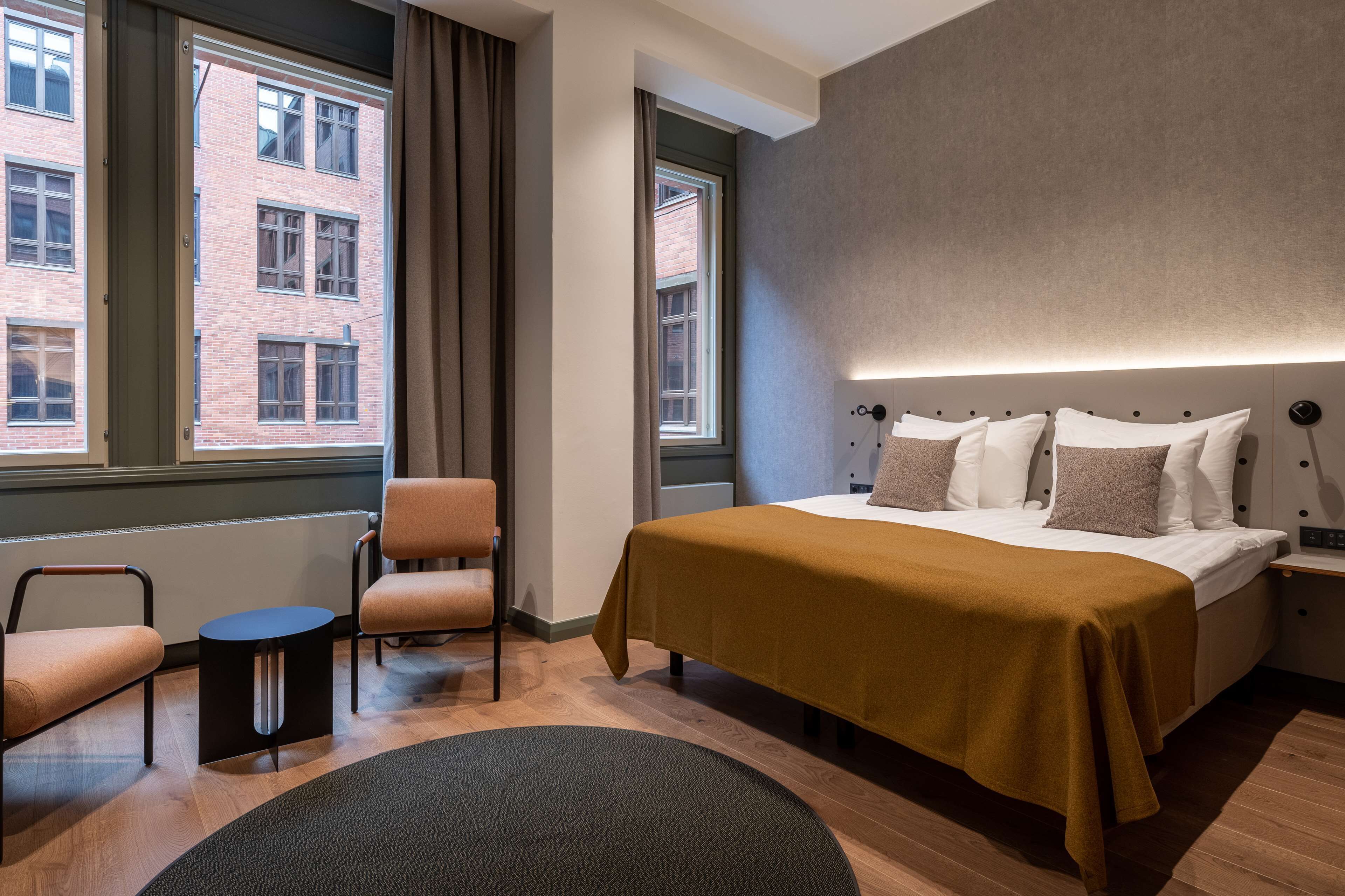 Une chambre d’hôtel moderne avec un grand lit, une chaise, une petite table et une vue sur un immeuble en briques par les fenêtres.