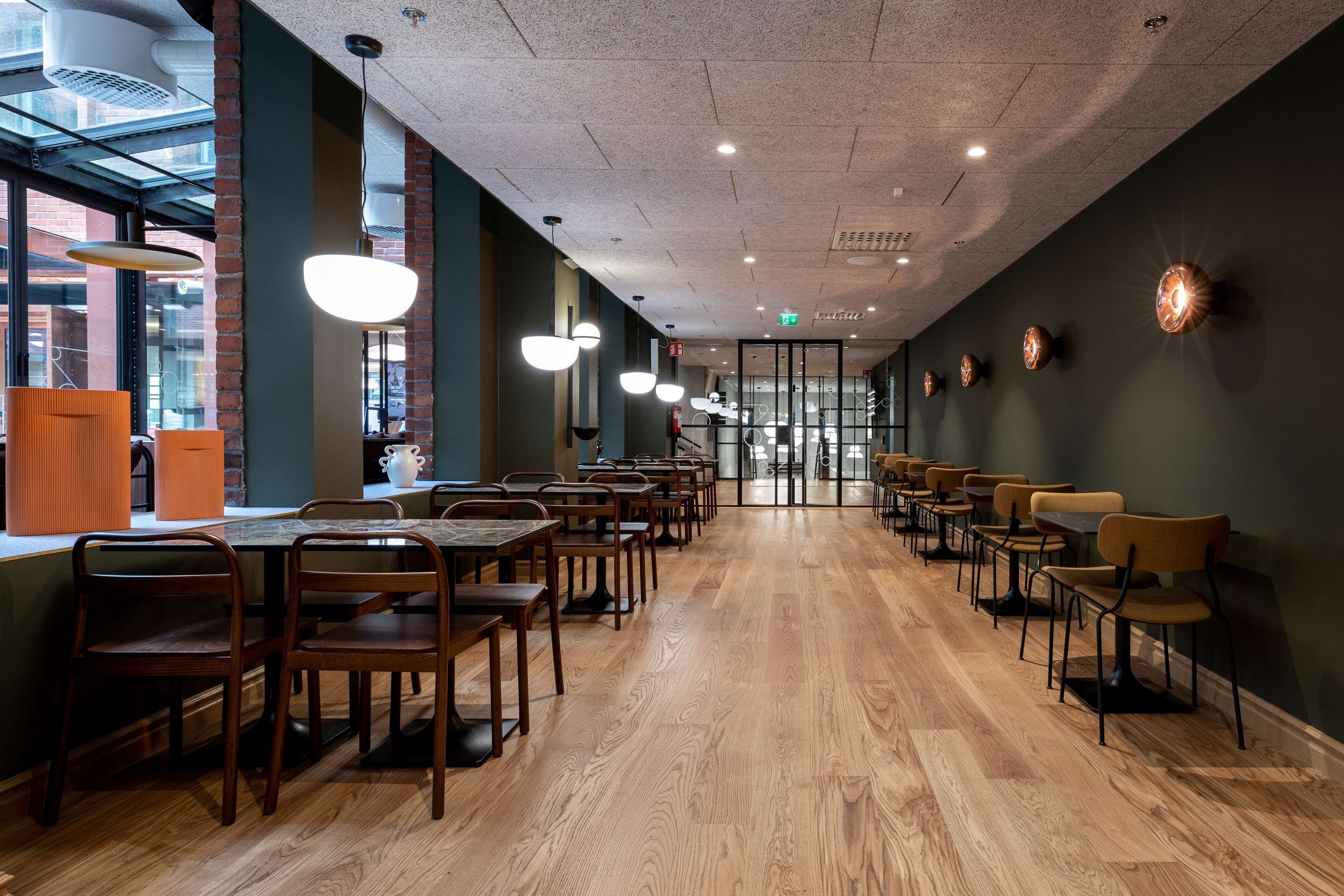 A spacious dining area with long wooden tables and chairs, modern pendant lighting, and a view through large windows.