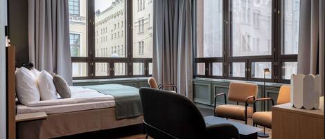 A modern hotel room with a bed, a desk, a chair, and large windows offering a view of the city.