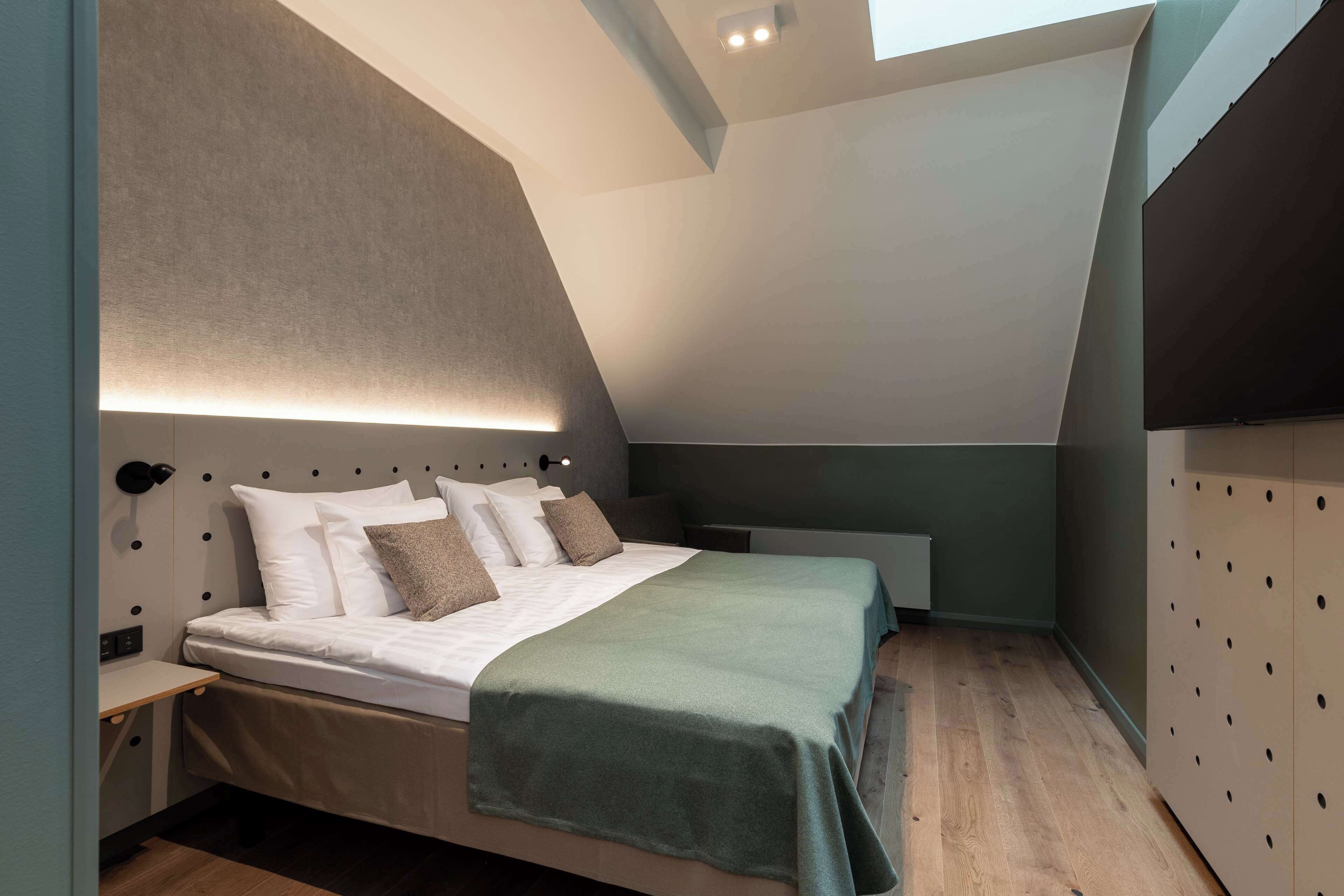 A modern bedroom with a sloped ceiling, a bed with white linens and a green throw, and a wall-mounted light fixture.
