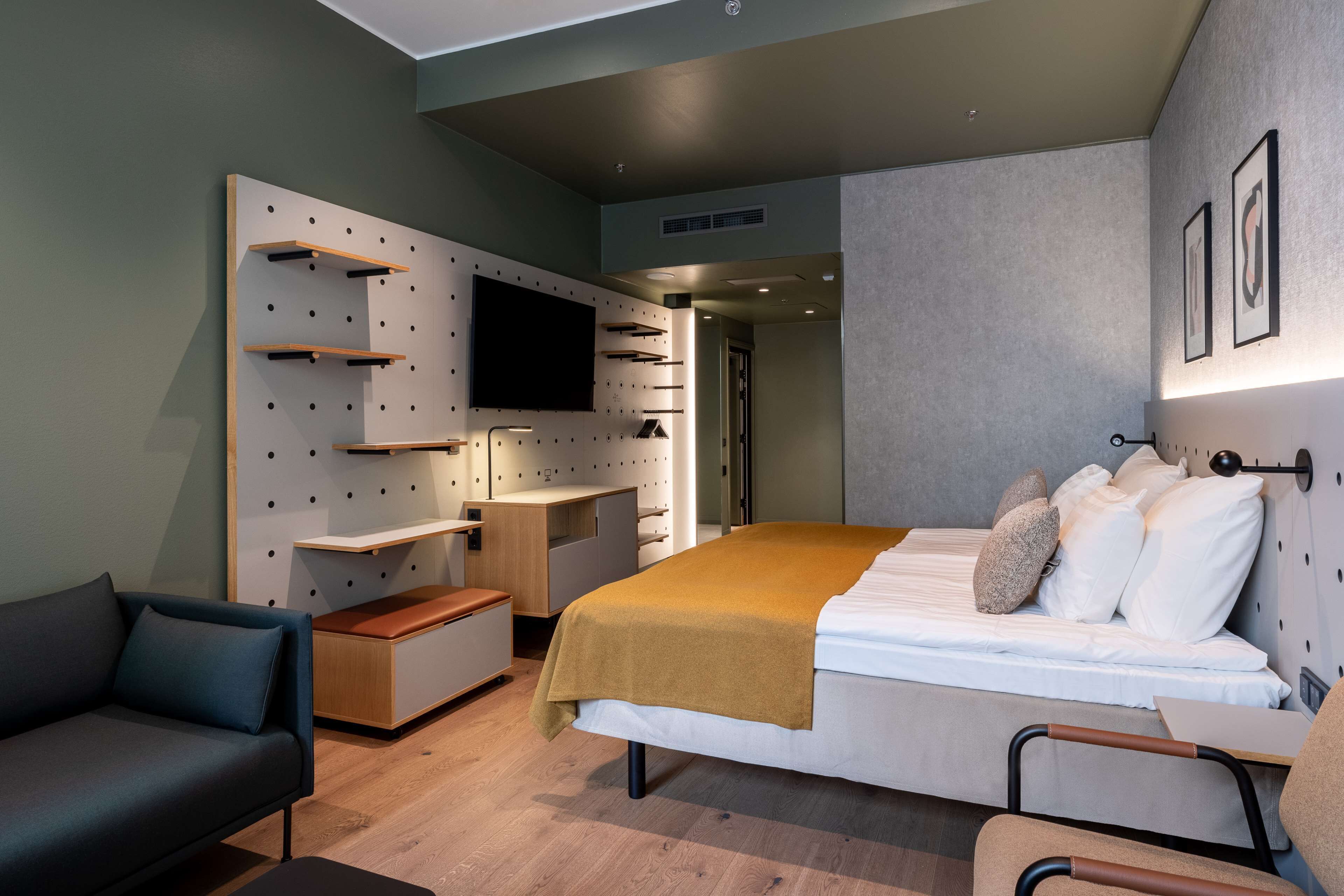 A modern hotel room with a bed, a sofa, a desk, and a TV mounted on the wall.