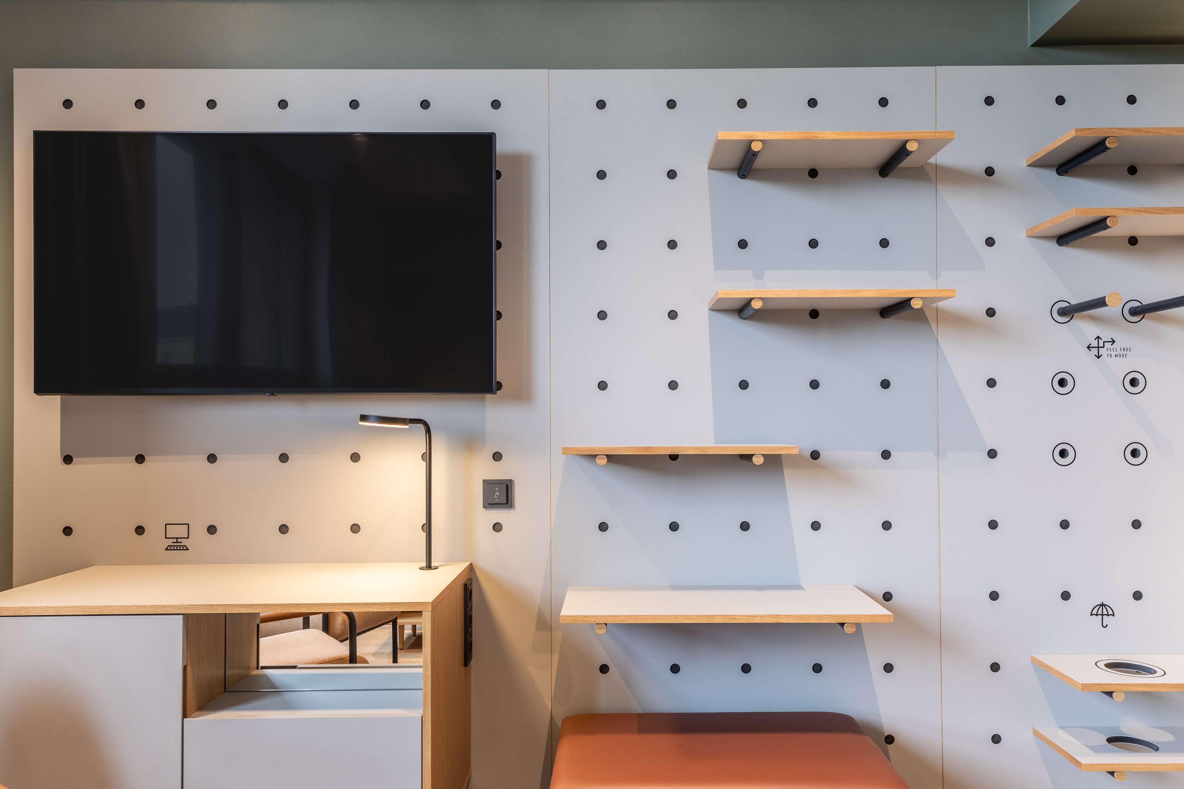 Una habitación moderna con un televisor de pantalla plana montado en una pared blanca, un escritorio con cajones y varios estantes de madera con ganchos.