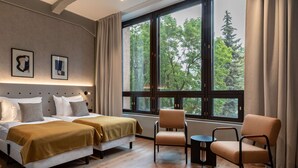Habitación de hotel con dos camas, una silla, una mesita pequeña y un ventanal con vistas a árboles.