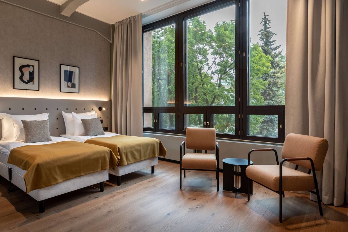 Une chambre d’hôtel avec deux lits, une chaise, une petite table et une grande fenêtre donnant sur des arbres.