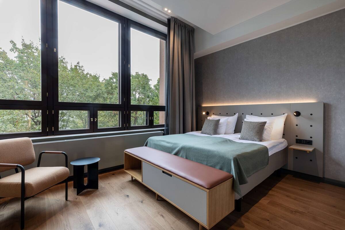 Une chambre à coucher moderne avec une grande fenêtre, un lit doté d’une tête de lit, un banc et une petite table.
