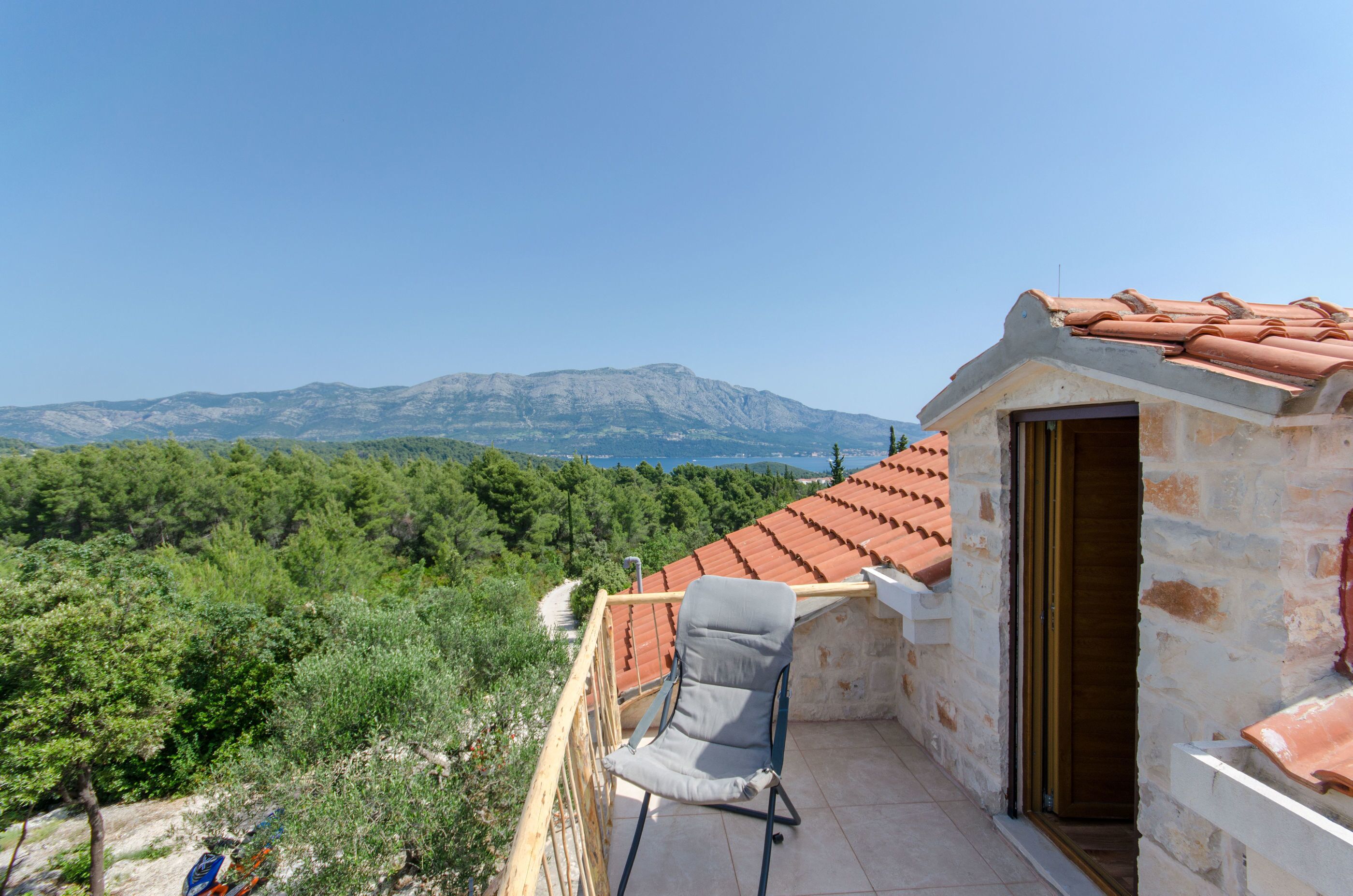 A balcony with a view of mountains, a chair, and a building with a tiled roof.