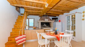 Un escalier en bois mène à une cuisine équipée d’une cheminée, d’une table à manger avec des chaises et d’un buffet blanc aux portes vitrées.