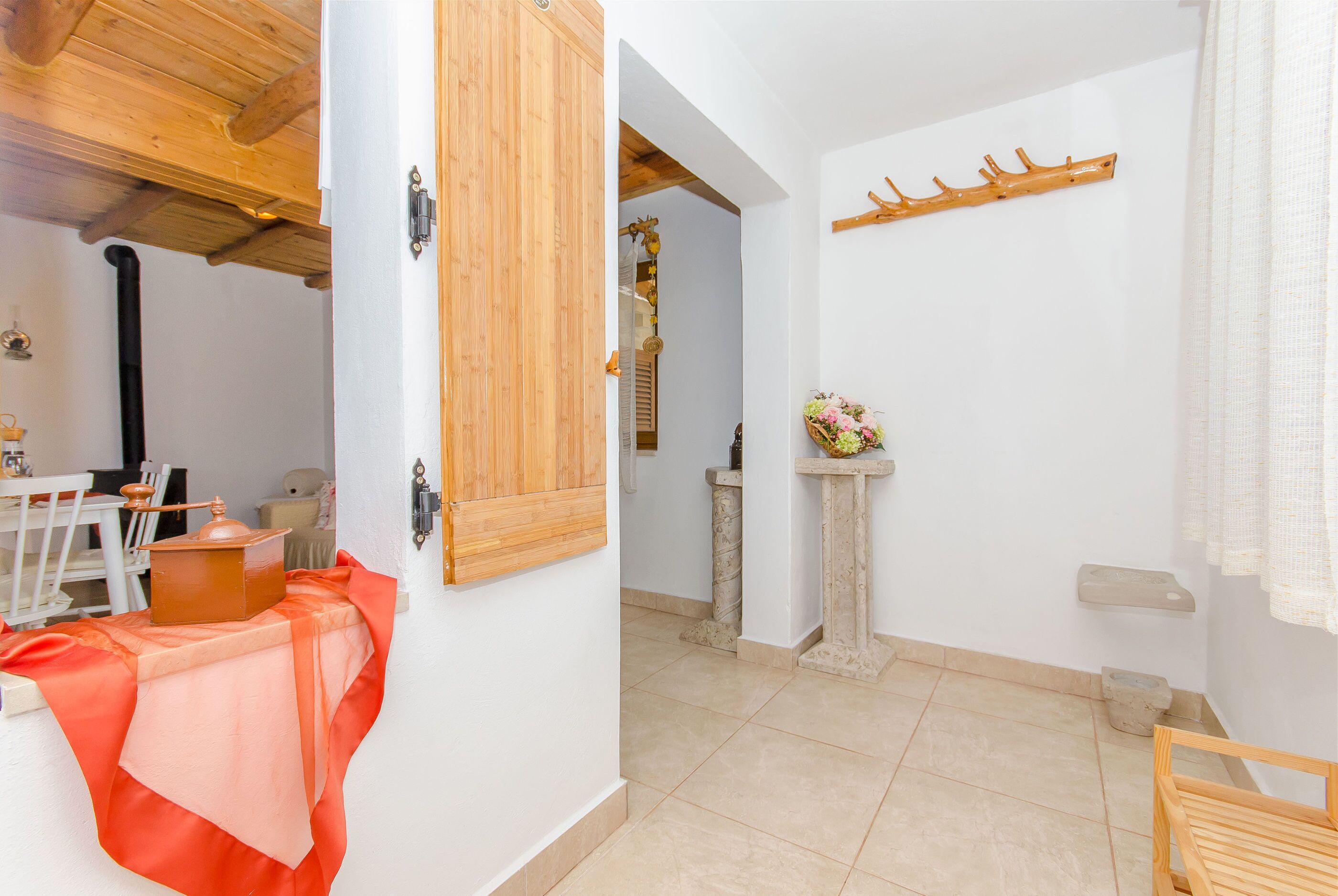 A room with a wooden ceiling, a table with a cloth, a wooden door, a wall-mounted coat rack, and a stone pedestal with a flower arrangement.