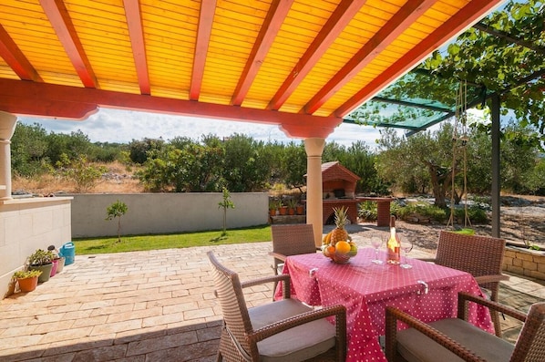 An outdoor patio with a brick floor, wicker furniture, and a table set with a fruit bowl and wine glasses. - Apartment Irena (Supetar)