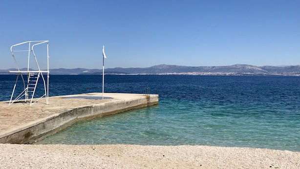 A concrete swimming platform with a ladder and a flagpole, overlooking a calm sea with distant mountains and a town. - Apartment Irena (Supetar)