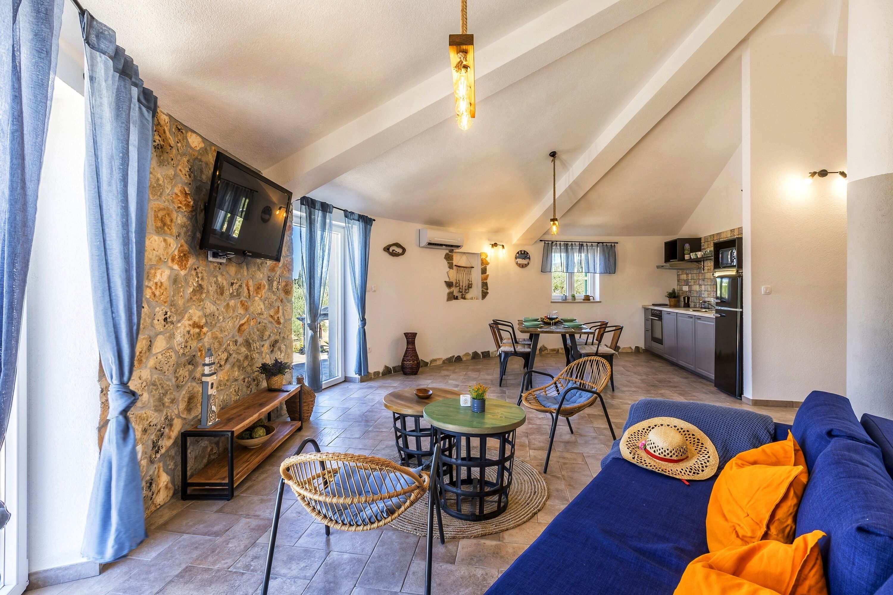 Un accogliente soggiorno con parete in pietra, televisione a schermo piatto, poltrone in vimini, un divano blu e una cucina sullo sfondo.