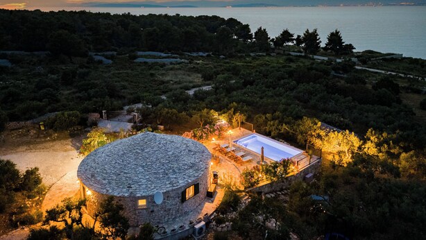 Un bâtiment circulaire en pierre, doté d’une piscine, entouré d’arbres et offrant une vue sur la mer.