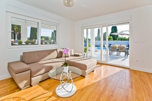 A modern living room with a beige sofa, a glass coffee table, and a view of an outdoor patio with a pool and furniture. - Villa Blazen (Bogomolje)