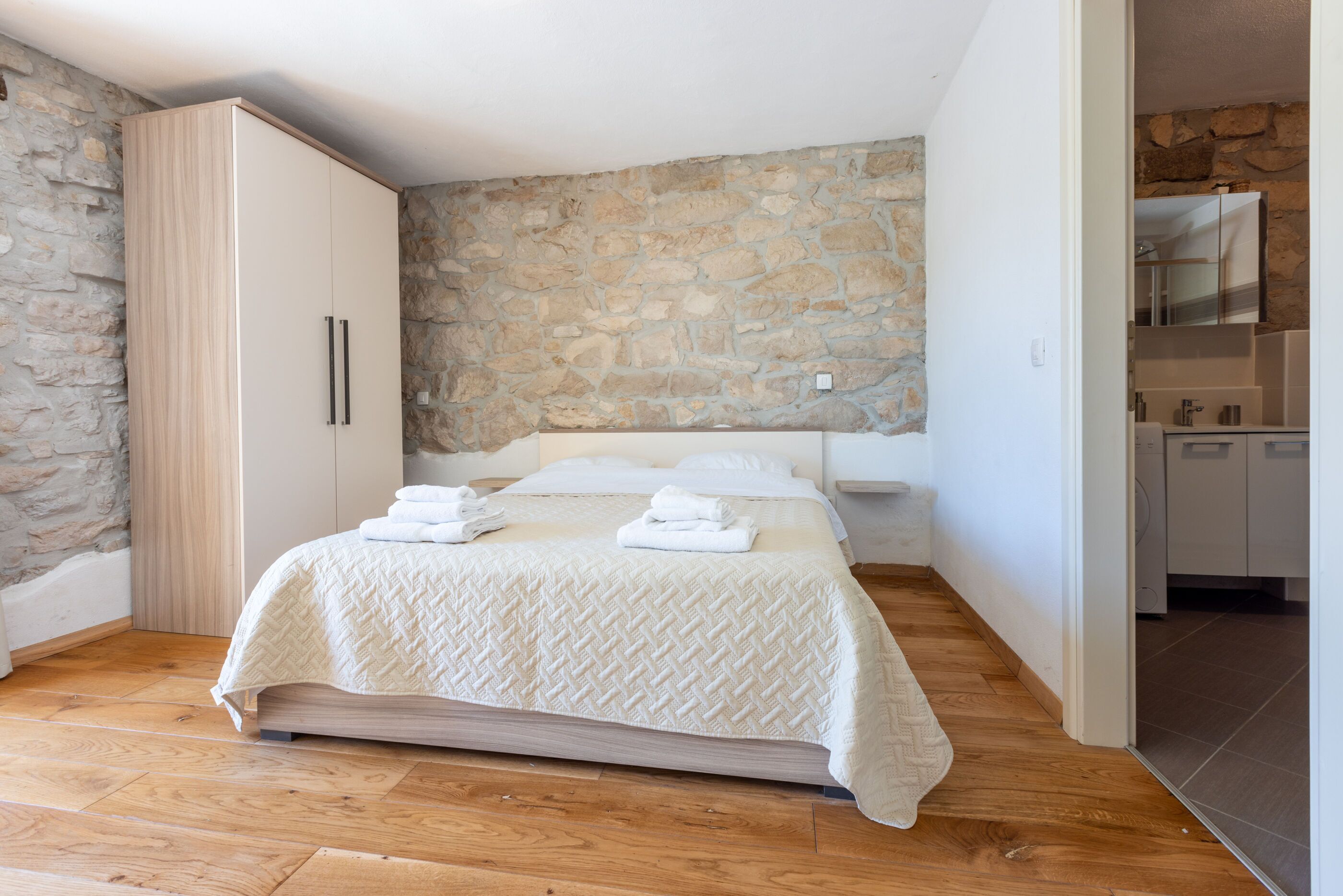 A bedroom with a stone wall, a wooden floor, a bed with a white quilt, and a wardrobe with a wooden finish.