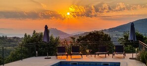 Ein Poolbereich mit Liegestühlen und Sonnenschirmen mit Blick auf eine malerische Landschaft, Hügel und einen Sonnenuntergang.