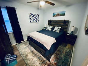 A bedroom with a bed, a ceiling fan, a nightstand, a lamp, a rug, and a painting on the wall.