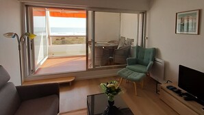 A modern living room with a sofa, a green armchair, a coffee table with a vase, a flat-screen TV, and a large window with a view of the outdoors. - SEAFRONT APARTMENT 60 M2 AND LOGGIA (Saint-Jean-de-Monts)