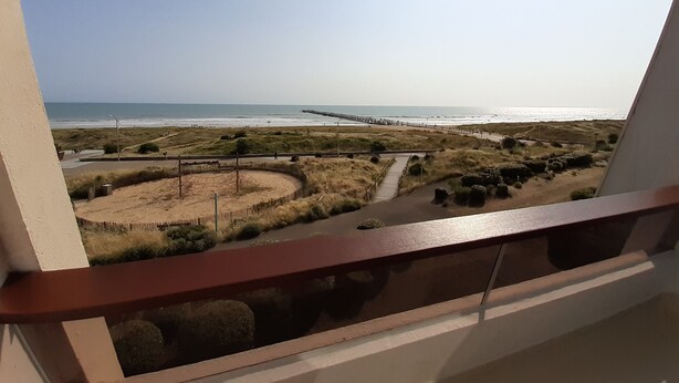 A view from a balcony overlooking a beach with a wooden walkway, grassy dunes, and a pier extending into the water. - SEAFRONT APARTMENT 60 M2 AND LOGGIA (Saint-Jean-de-Monts)