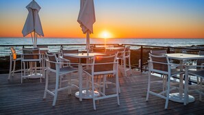 En træterrasse med hvide borde og stole, med udsigt over havet i solnedgang.