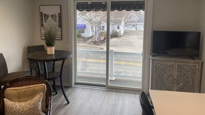 Une pièce comprenant une table à manger, des chaises, un téléviseur posé sur un meuble, une plante en pot et une porte-fenêtre coulissante donnant sur une vue d’une rue et de maisons.