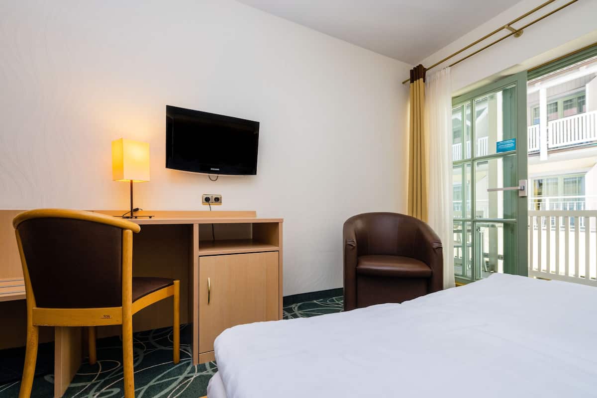 A hotel room with a bed, a desk, a chair, a television, and a view of a building with balconies.