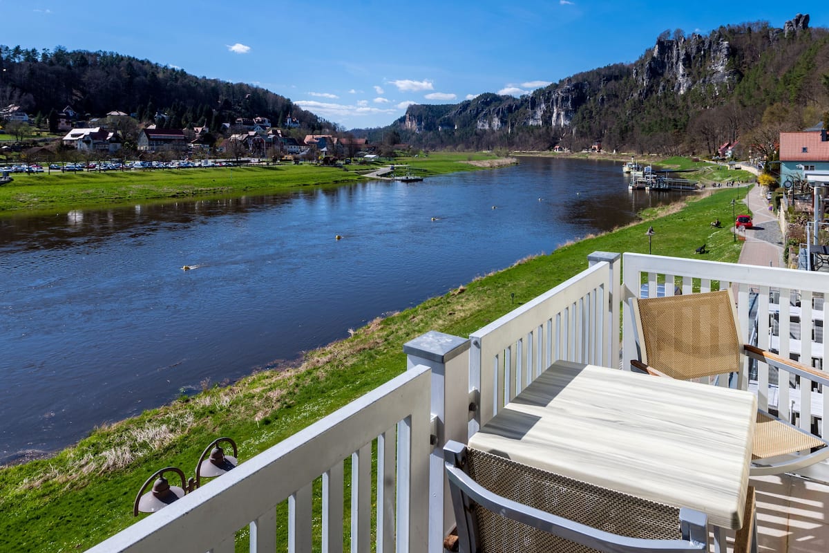 A riverside view from a balcony with a table and chairs, overlooking a river, a path, and a town with houses and a mountain in the background.