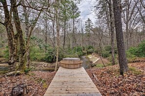 Una terraza de madera que lleva a un jacuzzi circular en un área boscosa con un arroyo.