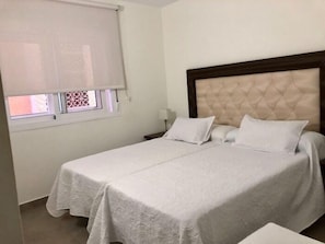 A room with two beds, a window with a blind, a nightstand, and a wall-mounted lamp.