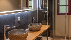 Une salle de bain avec deux vasques en pierre, un meuble-lavabo en bois, une cabine de douche en verre et un miroir avec un bandeau lumineux.