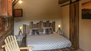 Une chambre avec une tête de lit en bois, un lit recouvert d’une literie à motifs, deux lampes de chevet, une chaise en bois avec un coussin moelleux et un plancher en bois.
