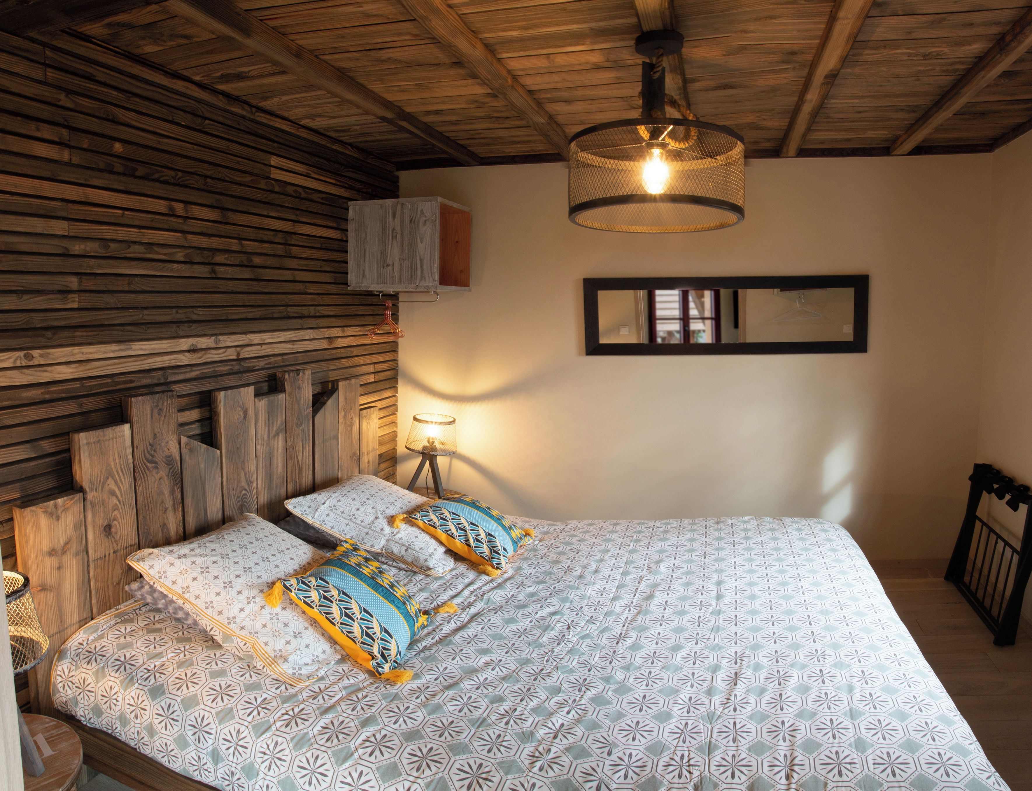 Een slaapkamer met een houten hoofdeinde, een bed met beddengoed met een patroon, een hanglamp, een nachtkastje met een lamp en een ingelijst schilderij aan de muur.