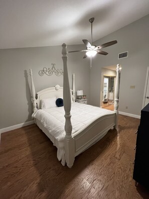 A four-poster bed with a ceiling fan, a nightstand, and a mirror in a bedroom with hardwood floors and light gray walls. - Spacious 2BD 2BA FreeWiFi Parking 12 mi - AT&T Stadium, Globe Life Field, TLive! (Arlington)