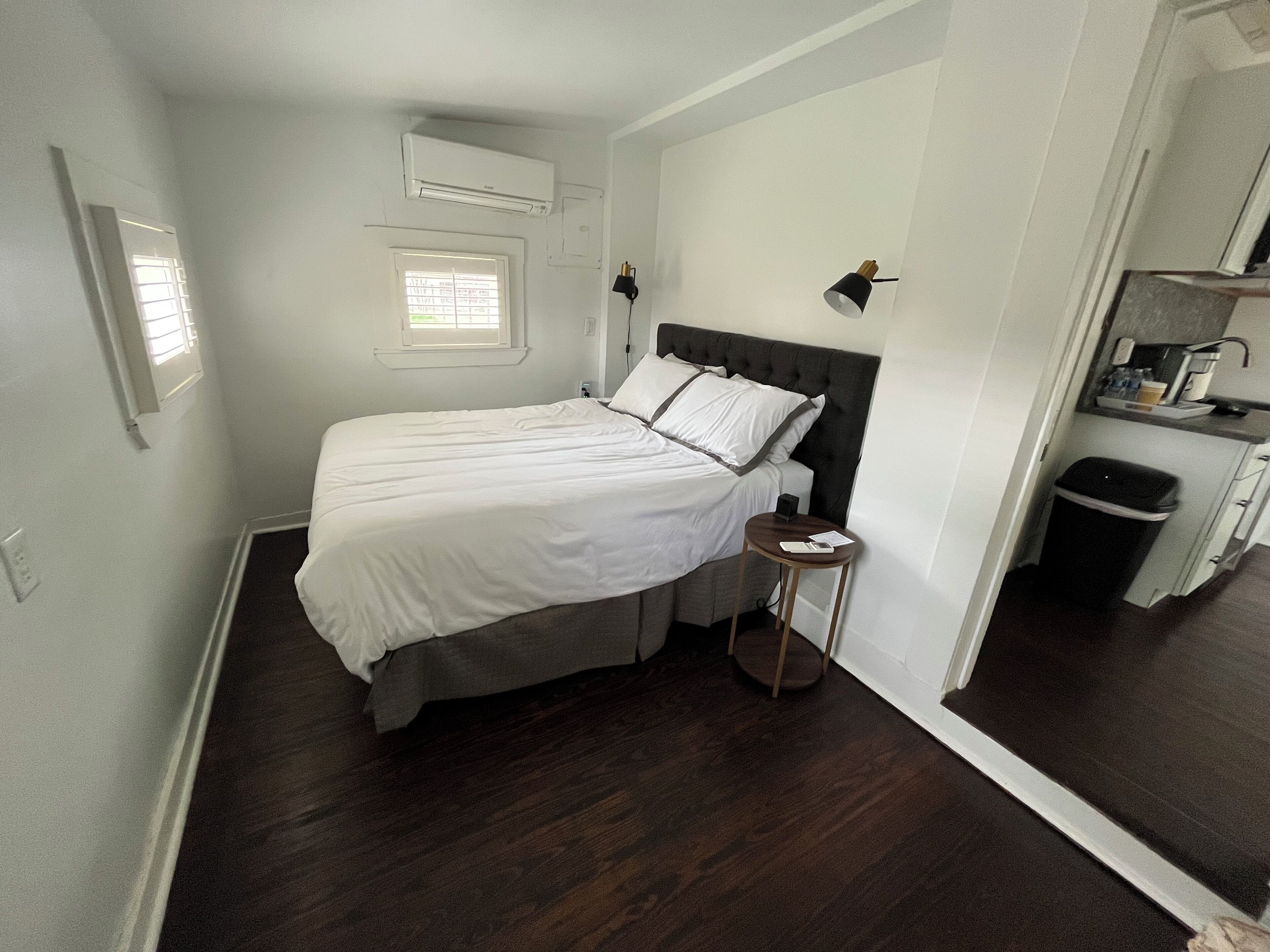 A bedroom with a bed, bedside table, and a small stool. There is a window with blinds, an air conditioning unit, and a door leading to a kitchenette.